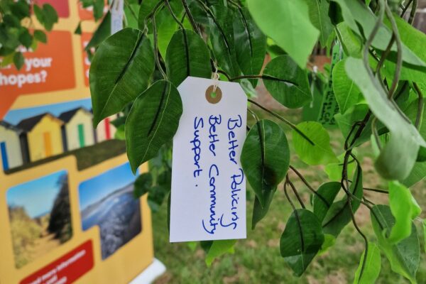 A tag hanging from a tree with a handwritten note on of what people want to see. Words on tag say - Better policing and better community support