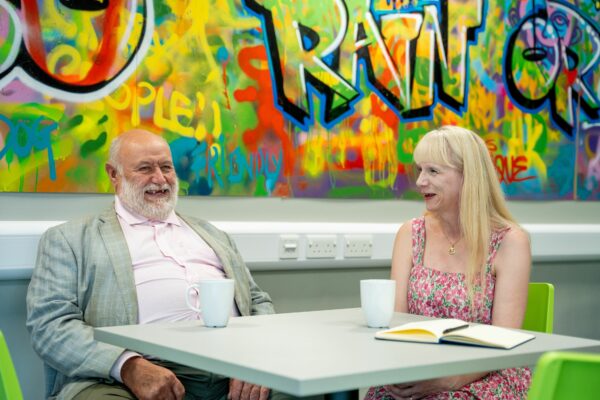 Man and lady sat at a table chatting. Having a cuppa together. There are two cups on the table and a colourful wall behind them. The couple are smiling and laughing.