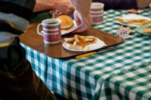 Image of a brown tray placed on a table with a green checkered table cloth on it. The tray has a mug of coffee on it and a plate with toast on it and a person's hand is removing a plate with a sausage sandwich on it.