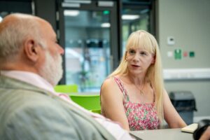 Lady with long blonde, straight hair and a fringe wearing a summer dress is sat down at a table chatting to a gentleman who is slightly blurred and to the left side of the image