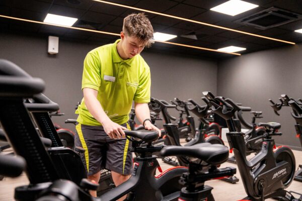 Image of leisure attendant wearing a lime green t-shirt working in a gym checking on exercise bikes