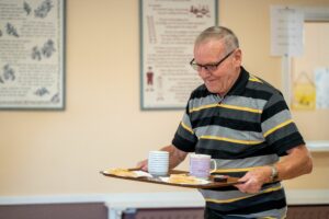 Image of an elderly gentleman carrying a tray with 2 mugs and plates of food on it. He is wearing glasses and a colourful stripey polo top.