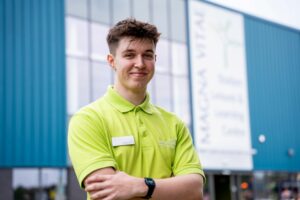 Close up of a young male leisure attendant in lime green t-shirt, stood outside of the building, looking at the camera and smiling.